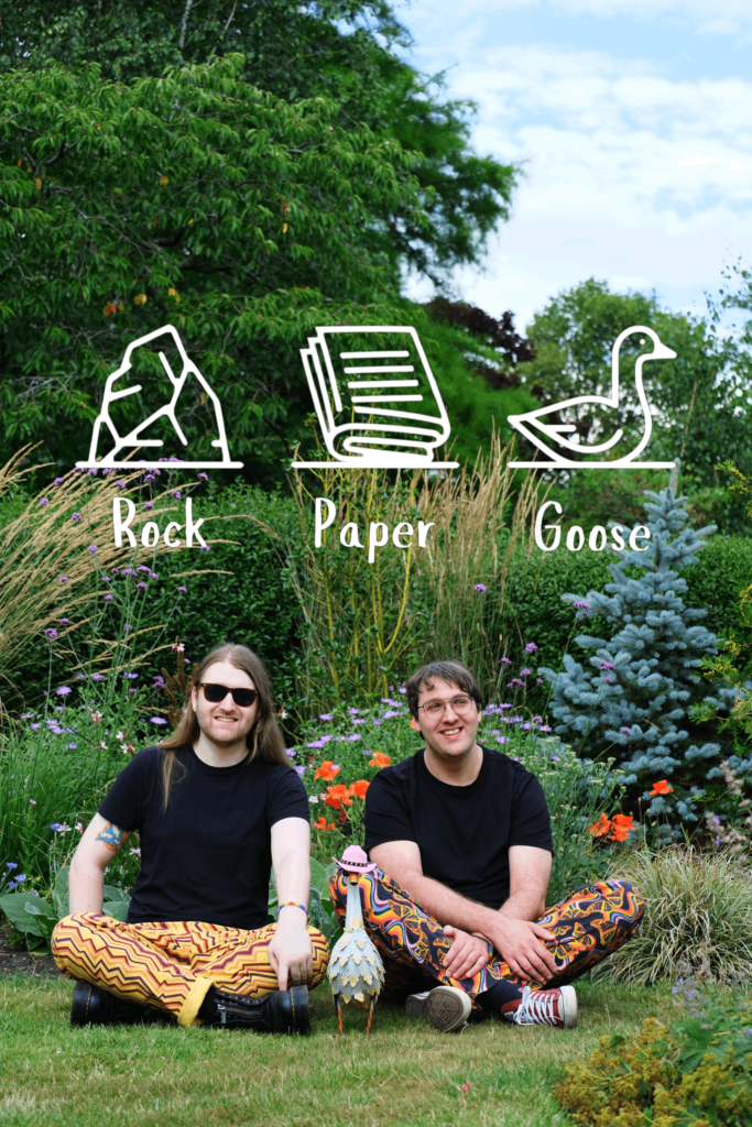 Nat & Olly from Rock Paper Goose sit among some lovely flowers and bushes. The Rock Paper Goose logo is emblazoned above. They're wearing some delightfully jazzy trousers and are accompanied by a small metal goose wearing a pink cowboy hat.